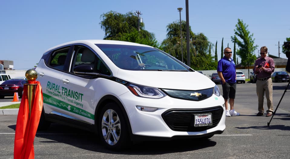 Photo of an electric Chevy Bolt car. The car has a wrap indicating it is part of the Rural Transit program sponsored by Measure C. It is a zero emissions vehicle.